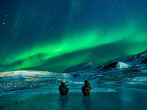 Two people sitting on ice and looking at polar lights.