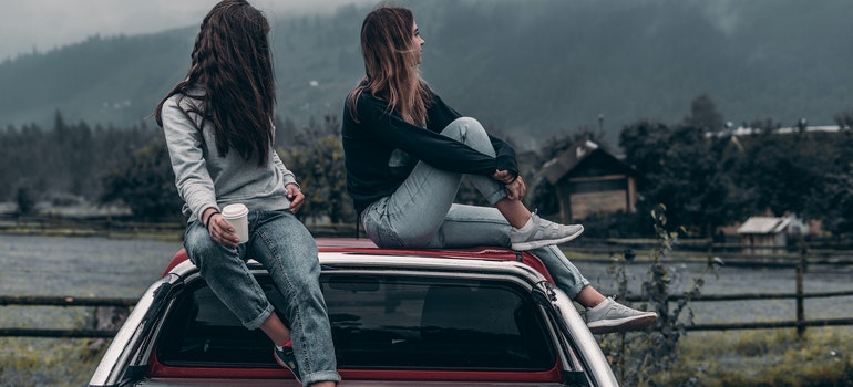 Two young women sitting on the roof of their car and looking at the distance.