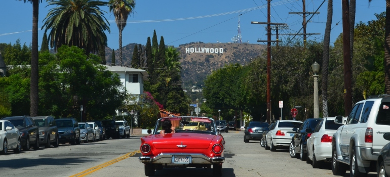 A man in a red car driving towards a hill with a Hollywood.