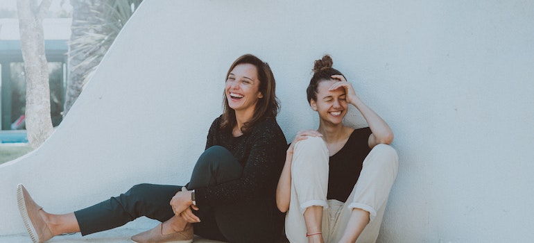 Two young women sitting with their backs on a wall and smiling.