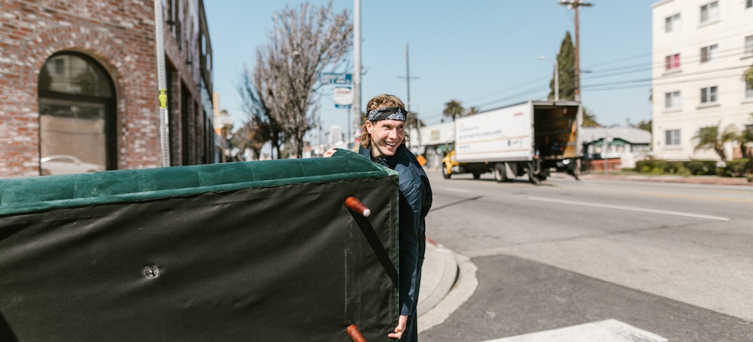 A man working for cross country moving companies Kansas smiling while holding a green couch in the street.
