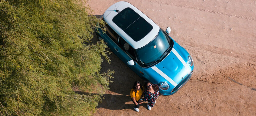 Two women photographed from air standing besides a blue car.