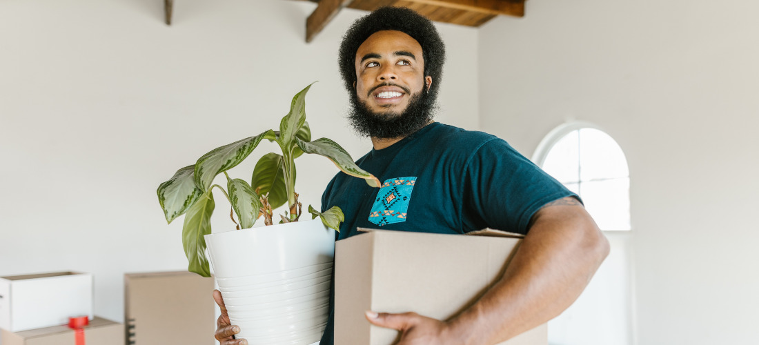 A guy who is working for long distance moving companies Virginia smiling while holding a box and a plant