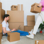 A man is on the floor typing something on his laptop while a woman is walking around carrying boxes