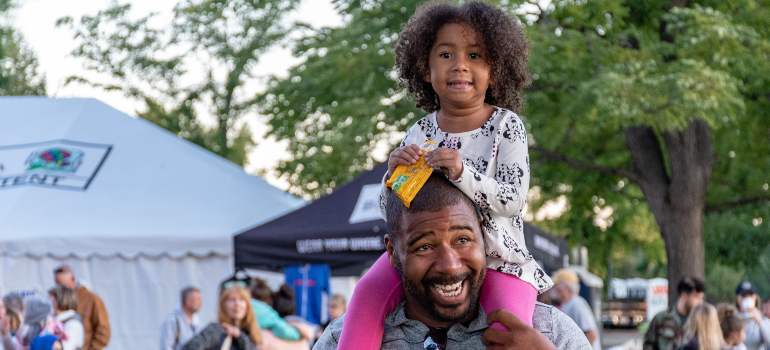 A girl is sitting on her father's shoulder while he's smiling.