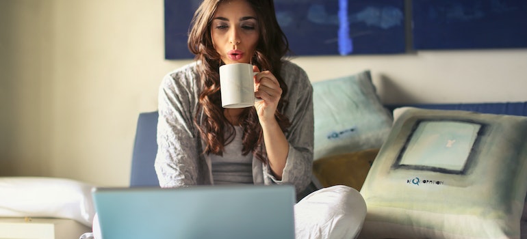 A woman on her bed drinking coffee and working on her laptop