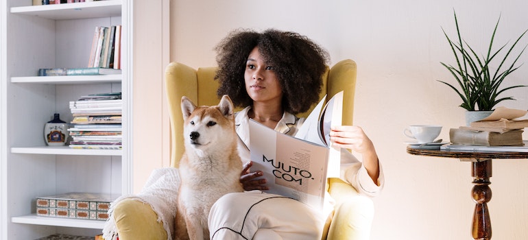 A woman sitting on a yellow chair with her Shiba Inu dog reading a magazine