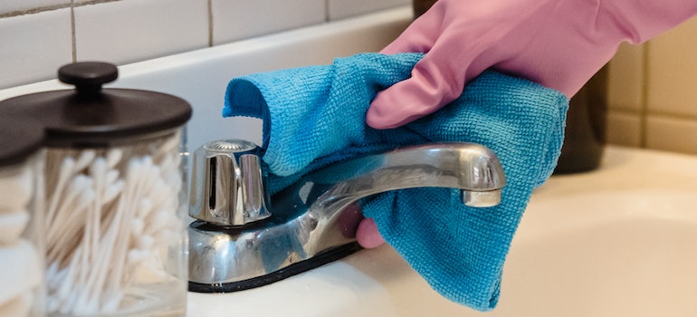 A person in pink gloves wiping a silver faucet with a blue damp microfiber cloth after reading a guide to deep cleaning your new home