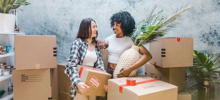 two women packing boxes for a cross country move with one of them holding a huge white ratan vase with palm leaves