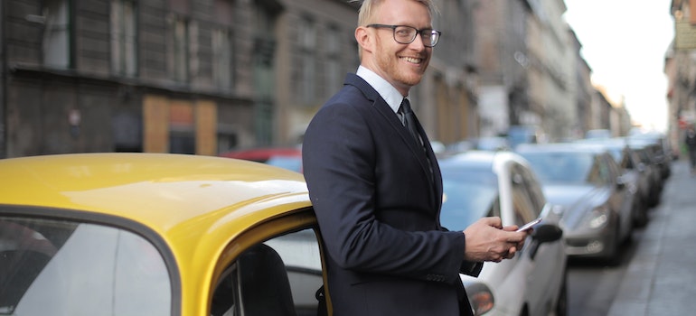 A man in a suit with glasses standing next to a yellow vintage car 