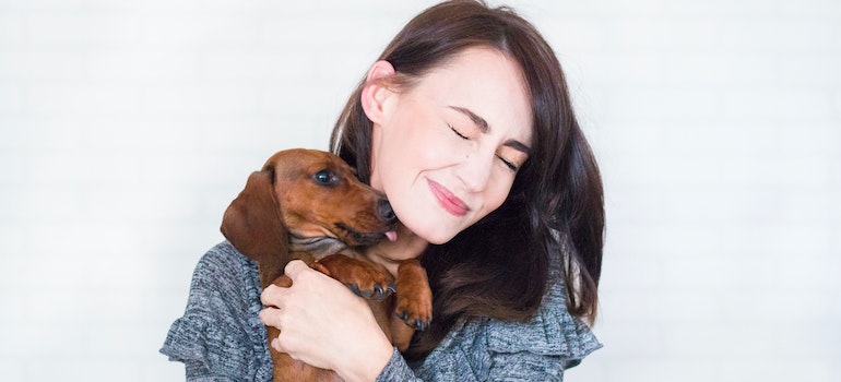 A woman with brown hair and a grey sweater carrying and hugging a brown dachshund dog