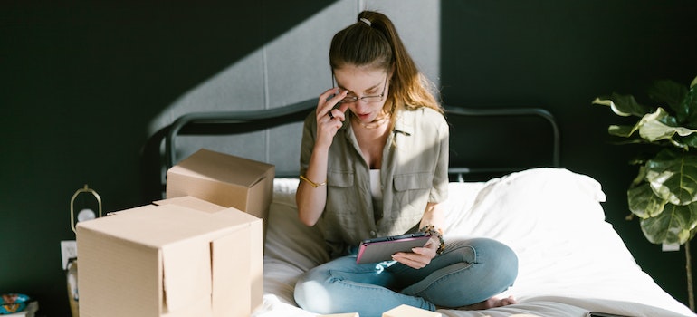Woman with glasses and a high ponytail looking at a tablet while sitting in bed with a bunch of cardboard boxes around her