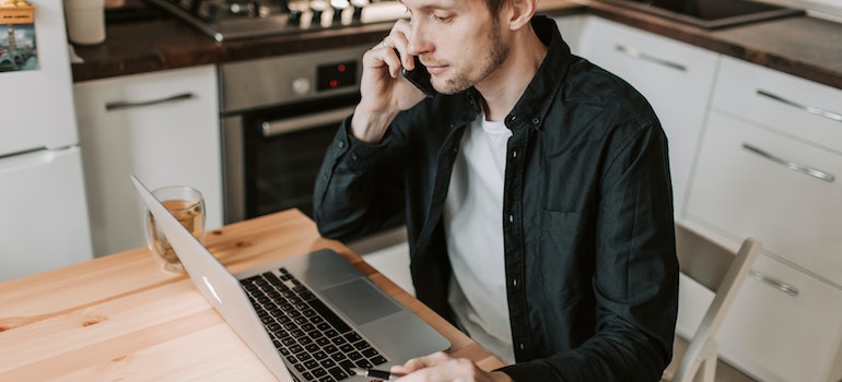 Man in a kitchen talking on a phone with insurance companies while looking at a grey Macbook