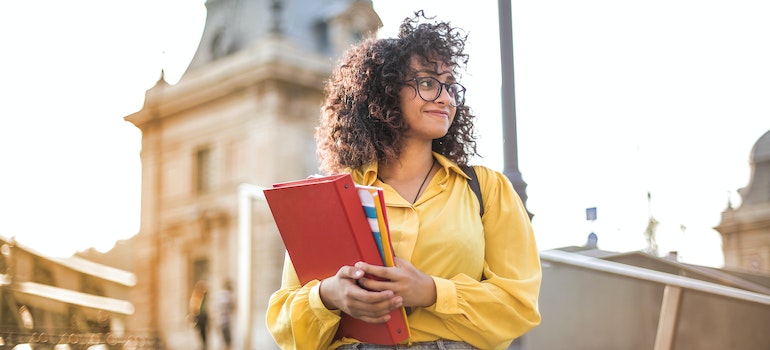 Woman in a yellow blouse with curly hair and glasses holding notebooks and a binder in her arms in front of a blurred college building