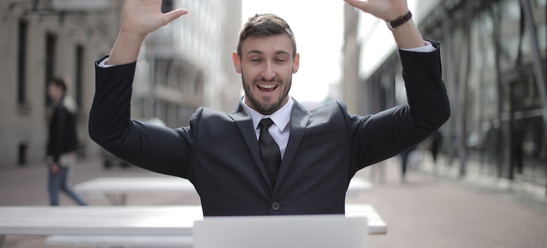Man in a black suit looking at a laptop with both hands in the air