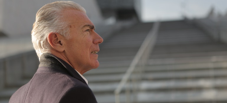 An old man with grey hair and a black coat standing in front of blurred stairs in the background