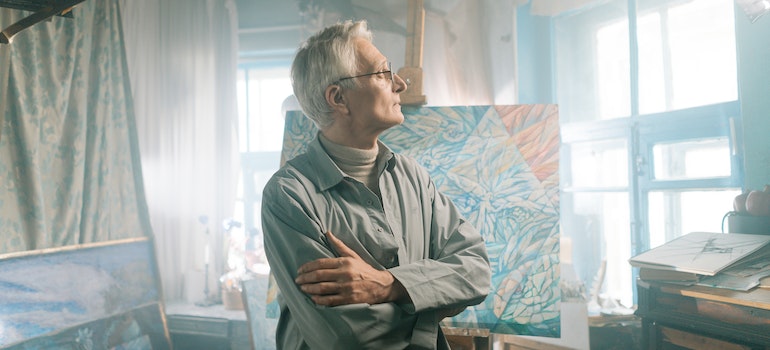 An older artist standing in front of his paintings in a studio after moving from Oklahoma City to Dallas