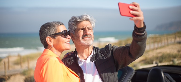 Two older people taking a selfie with a red phone in front of a sandy beach in Florida