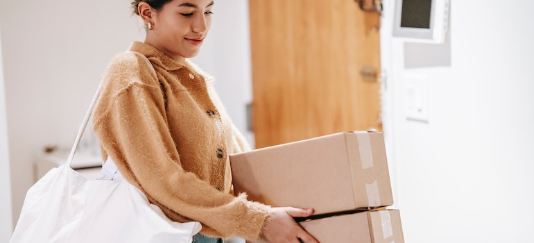 Woman in a slick bun and a brown shacket holding two cardboard boxes