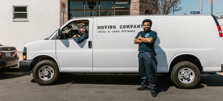 A man standing in front of a white moving van, while another man it sitting in it.