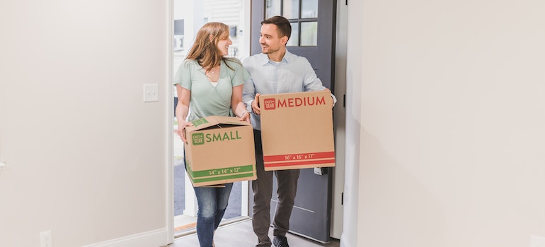 A couple carrying cardboard boxes entering their new home smiling after moving from New York to Maryland