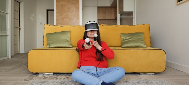 Woman in a red sweater sitting in front of a mustard couch with green satin pillows using a VR headset and wands