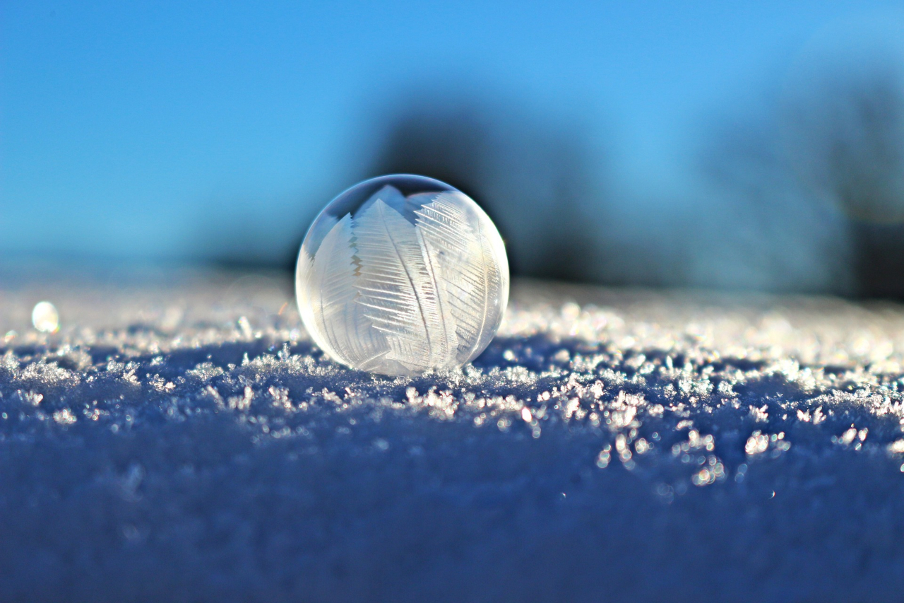 A frozen bubble on a snow-covered surface with ice crystals forming on its surface, set against a blurred background with a hint of sunlight..
