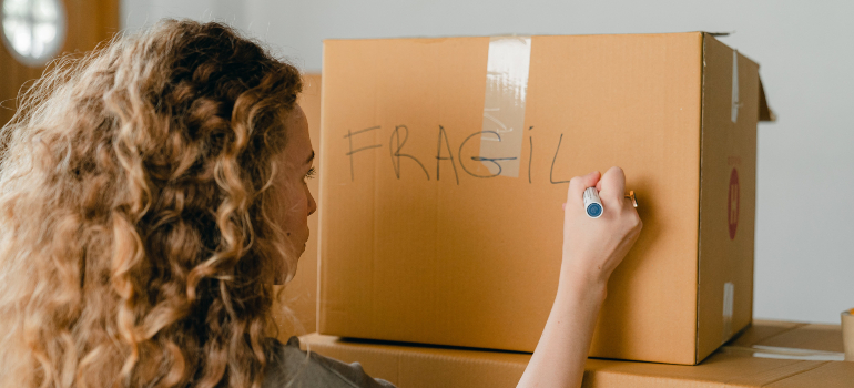 A woman marking a cardboard box with the word "FRAGILE" as she prepares to pack your Christmas decorations for an international move.