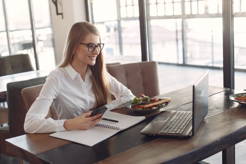Young cheerful businesswoman using smartphone and laptop in cafe during lunch time
