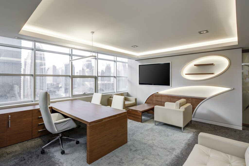 Gray Office Rolling Chair Near Brown Wooden Desk in Front of Flat Screen Tv on White Painted Wall
