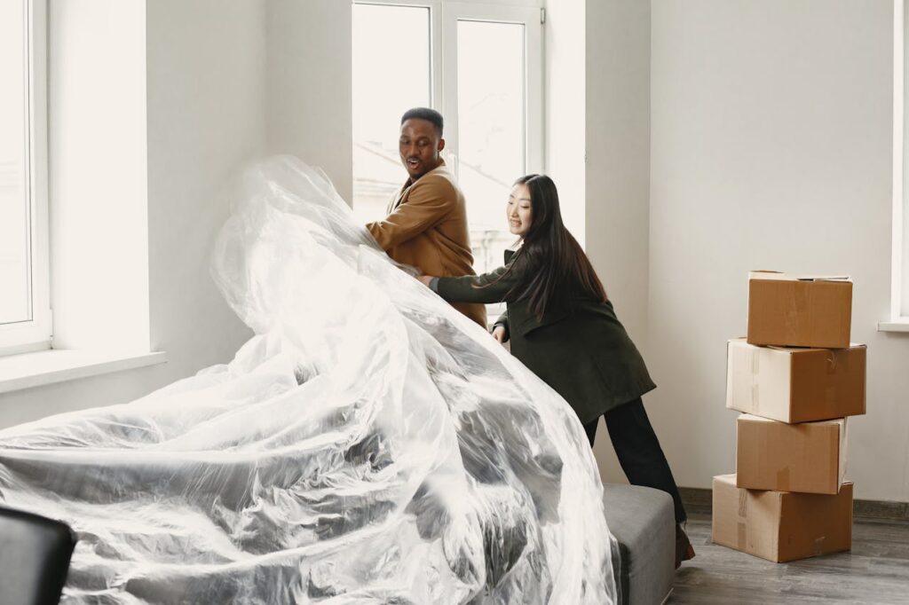 Man in Brown Coat Pulling the Plastic Cover on Couch with Woman in Black Coat
